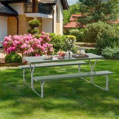 a picnic table with two benches in the grass near flowers and bushes on a sunny day