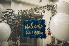 a welcome sign hanging from the side of a building next to balloons and greenery