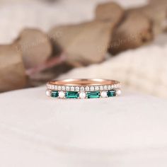 an emerald and diamond band ring sitting on top of a white stone surface with rocks in the background