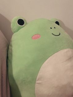 a green frog pillow sitting on top of a bed next to a white wall with black eyes