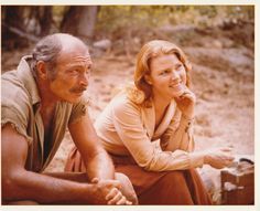 an older man and woman sitting next to each other in front of a forest area