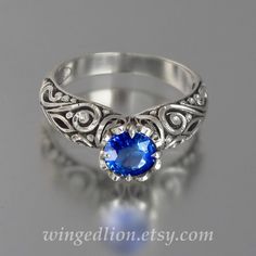 "BEATRICE ring is made to order in sterling silver in the size specified by the customer. The ring's shank is adorned with intricate scroll carvings, and the ornamented crown is prong set with a 6mm round lab-created Blue Sapphire (about 1 Ct, eye clean). There are four 1.2mm round natural white sapphires on the crown's sides. The crown is about 8mm high (including the stone) and 7.5mm wide. The widest part of the shank is 5.9mm. The ring can be made in sizes from 4 to 9. The ring is oxidized to