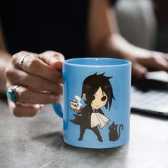 a person holding a coffee mug with an anime character on it, sitting in front of a laptop