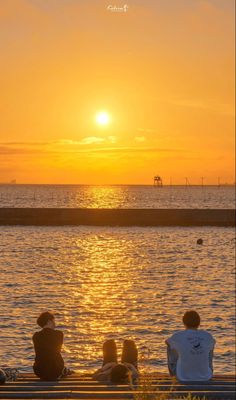 three people are sitting on a bench watching the sun go down over the water at sunset