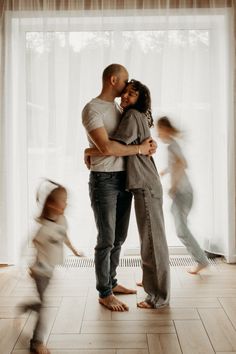 a man and woman are hugging in front of a window with children running around them