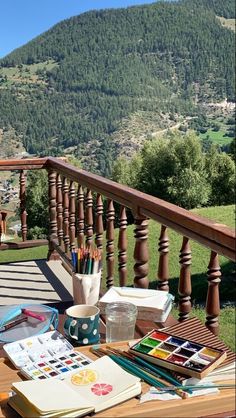 a wooden table topped with lots of art supplies next to a mountain side area covered in greenery