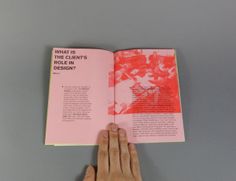 a hand holding an open book on top of a gray table next to a white and red object