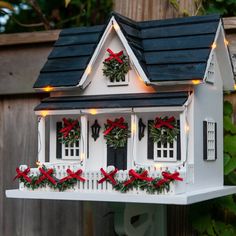 a white birdhouse with christmas decorations and lights on it's roof in front of a wooden fence