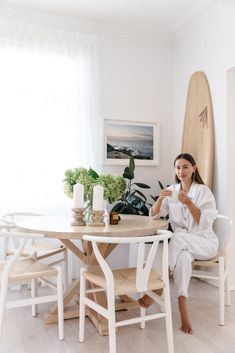 a woman sitting at a table with a surfboard in the background and plants behind her