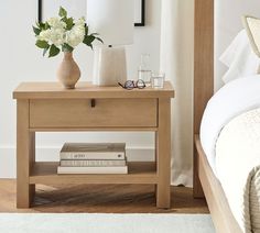 a nightstand with two books on it next to a bed and flowers in a vase