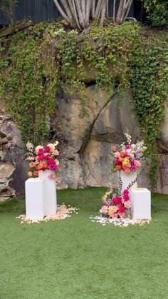 two white pedestals with flowers on them in front of a rock wall and green grass