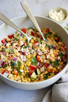 a bowl filled with corn and vegetables next to two spoons on a white surface