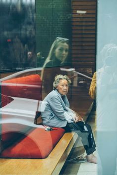 an older woman sitting on a bench in front of a glass window looking at her reflection