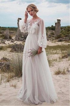 a woman standing in the sand wearing a white dress with sheer sleeves and long sleeves