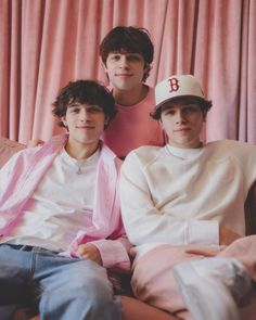 three young men sitting next to each other in front of a pink curtain, one wearing a baseball cap