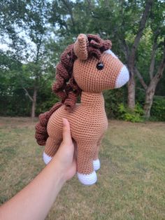 a hand holding up a crocheted stuffed horse in the grass with trees in the background