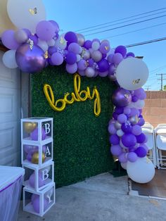 balloon arch decorated with purple and white balloons that spell out the word baby in front of a green wall