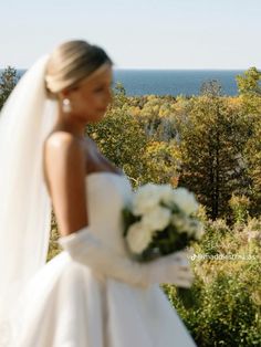 a woman in a white wedding dress holding a bouquet and looking out over the ocean