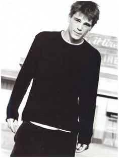 a young man standing in front of a building wearing a black sweater and pants with his hands in his pockets