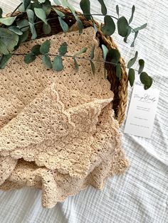 a crocheted blanket is in a woven basket on a bed with eucalyptus leaves