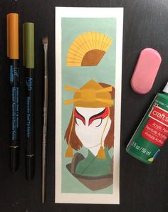 some pens and markers are sitting on a table next to an art print with a woman's face