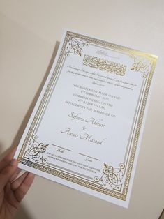 a person holding up a white and gold wedding card with arabic writing on it, in front of a beige wall