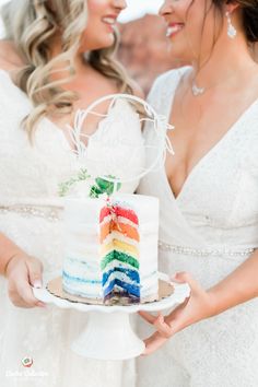 two women in white dresses holding a rainbow cake