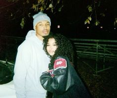 a man and woman standing next to each other in front of a fence at night