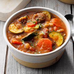 a white bowl filled with vegetable soup on top of a wooden table next to a spoon