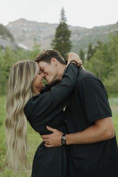 a man and woman embracing each other in front of some trees with mountains in the background