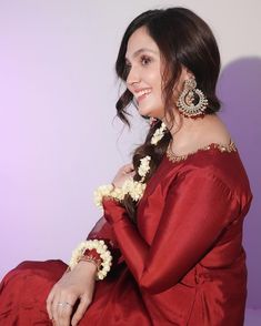 a woman in a red dress with flowers on her hair and earrings, sitting against a purple background