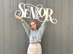 a woman holding up a sign that says senior in front of a gray wall with the word senior on it