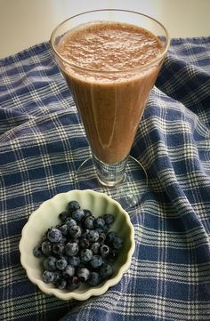 a bowl of blueberries next to a glass of smoothie on a checkered cloth