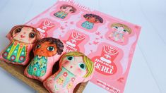 three clay dolls sitting on top of a wooden tray next to a pink paper with stickers