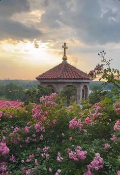 a cross on the top of a building surrounded by flowers and greenery at sunset