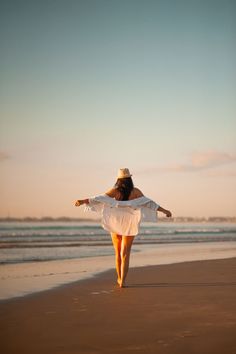 a woman walking on the beach with her arms spread out and wearing a white dress