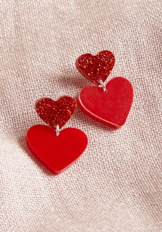 two red heart shaped earrings sitting on top of a pink cloth covered surface with glitter