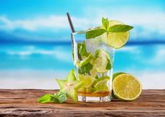 a glass filled with ice and limes on top of a wooden table next to the ocean