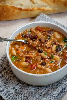 a white bowl filled with beans and meat next to a piece of bread on top of a blue towel