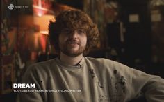 a man with curly hair wearing a sweater and looking at the camera while sitting in front of a wall