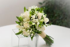 a bouquet of white flowers sits in a clear vase on a table with greenery