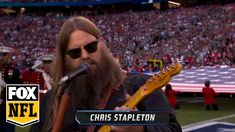 a man with long hair and sunglasses holding a guitar in front of an american flag