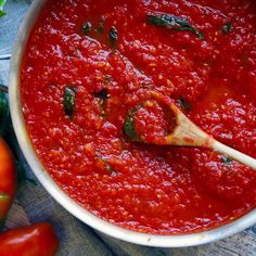 a bowl of marinara sauce with a wooden spoon in it next to tomatoes and basil