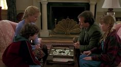 three people sitting around a table with a cake on it in front of a fireplace