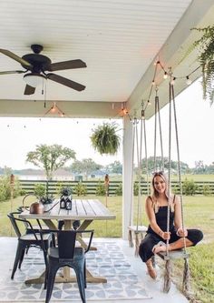 a woman sitting on a porch swing in front of a table with chairs and a ceiling fan