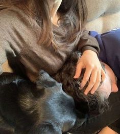 a woman laying on top of a couch next to a black dog and holding it's head