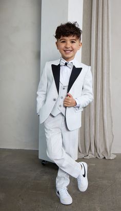 a young boy in a white suit and black bow tie standing next to a wall