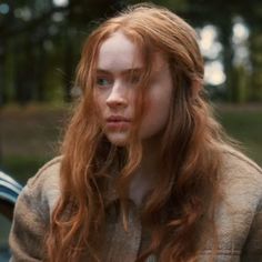 a woman with long red hair standing in front of a car and looking at the camera