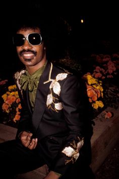 a man in a suit and sunglasses sitting on a ledge with flowers behind him at night