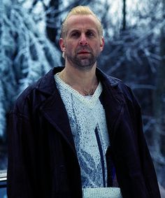 a man standing next to a car in front of trees with snow on the ground